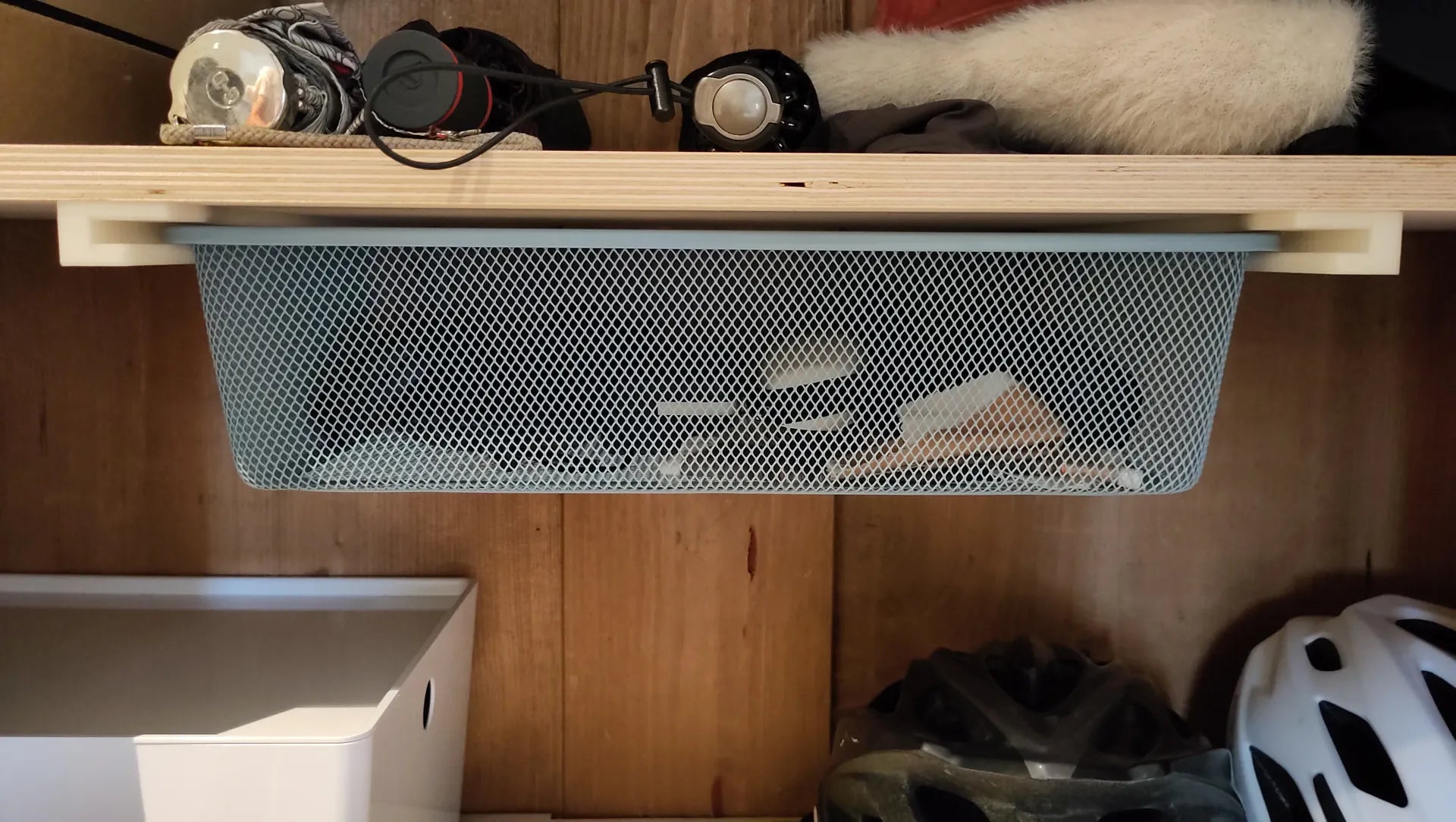 Metal mesh storage basket hanging on a wooden shelf with various items below.